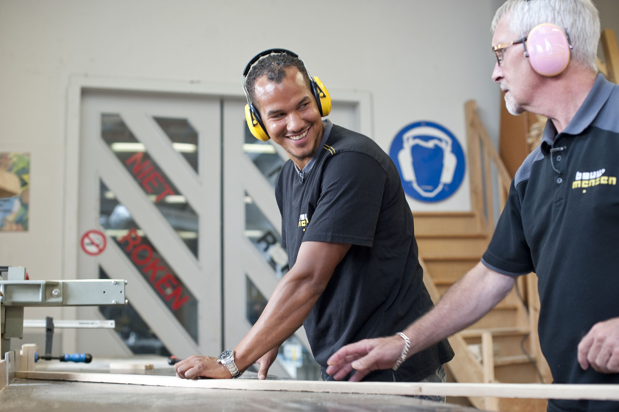 Twee timmerlieden dragen gehoorbescherming en overleggen tijdens het bewerken van hout in een werkplaats.
