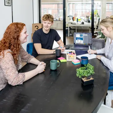 Drie studenten bespreken samen bij een tafel ideeën voor een welzijnsproject.
