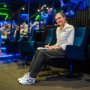 Medewerker evenementenorganisatie noteert tijdens briefing in zaal met aanwezigen op stoelen.