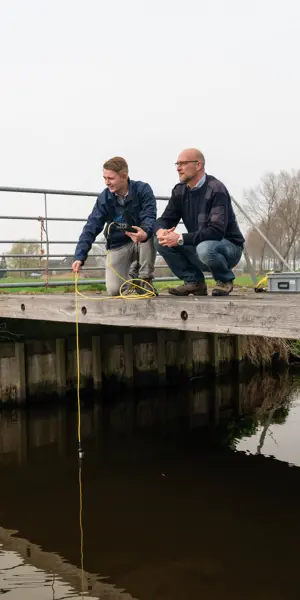 Twee mensen bekijken waterkwaliteit met technische meetapparatuur bij een houten brug over een waterloop