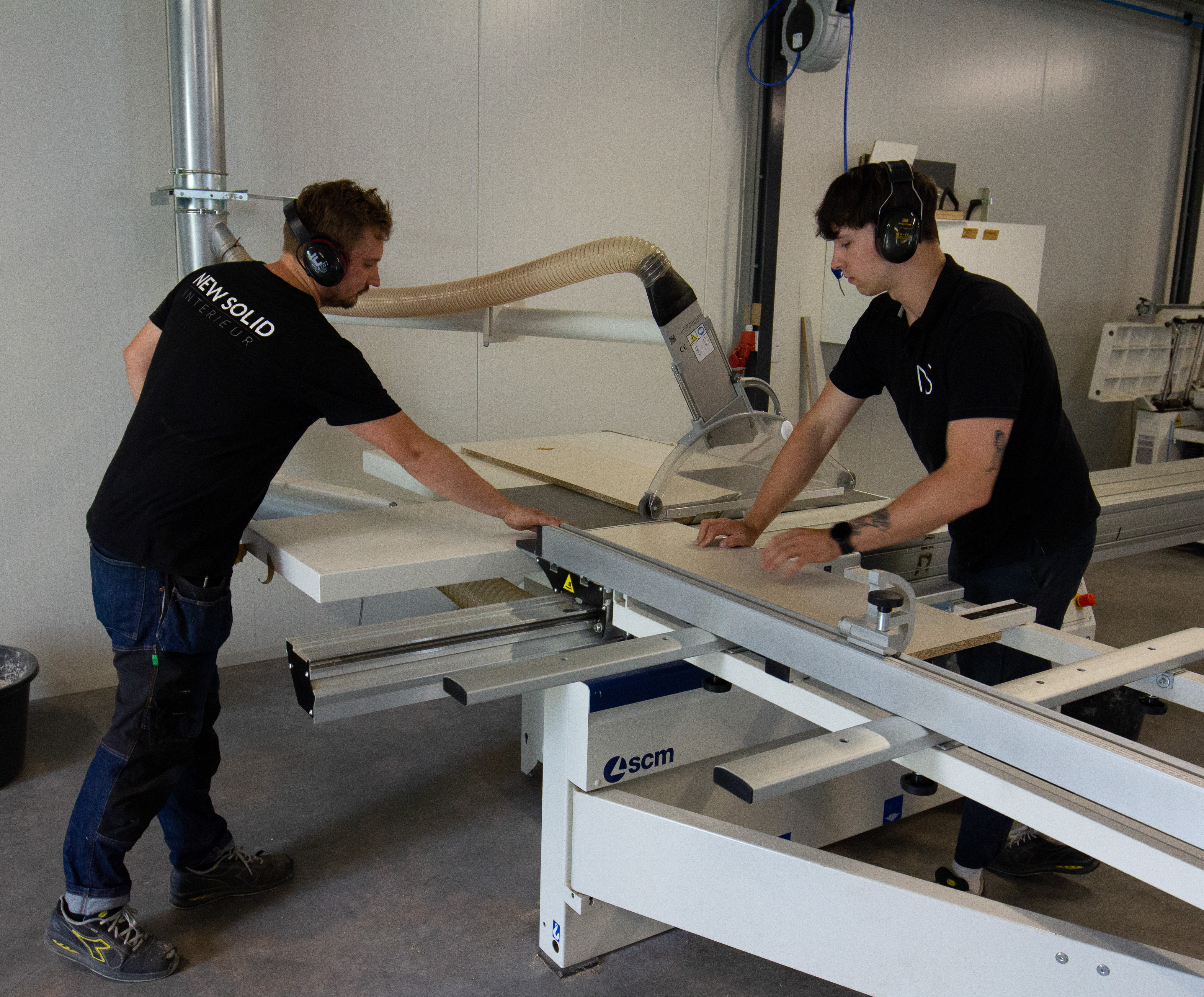 Twee meubelmakers bedienen samen een zaagmachine in een werkplaats voor houtbewerking.