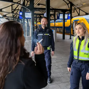Beveiligers in uniform spreken met een reiziger op een treinstation over veiligheid en ordehandhaving.