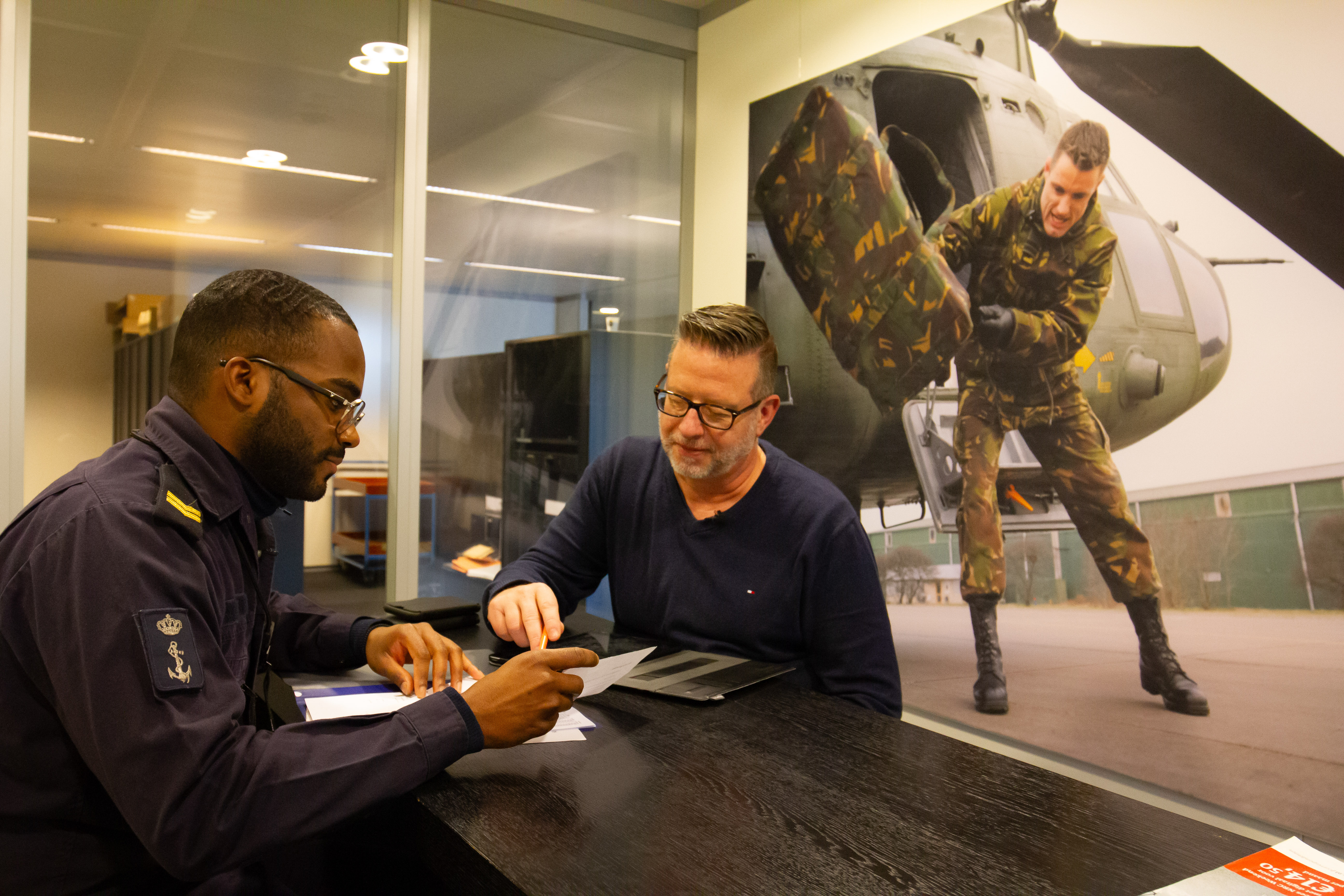 Twee personen overleggen aan tafel over documenten met een helikopter en militair op de achtergrond