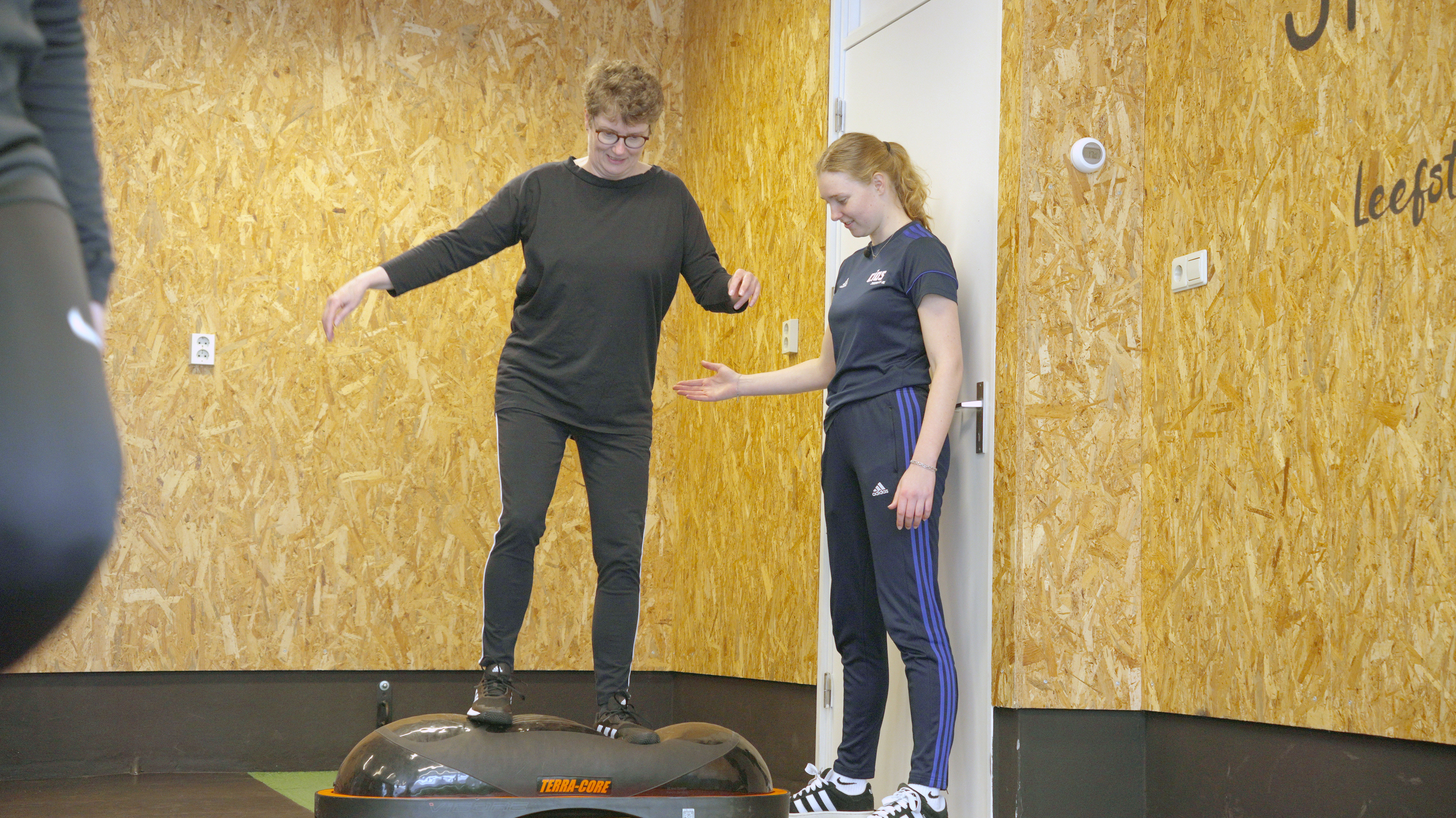 Begeleider ondersteunt student tijdens oefening op Balance Trainer in een sportomgeving