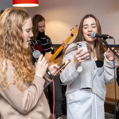 Jongeren oefenen zang en basgitaar in een oefenruimte voor podiumtechniek en entertainment.