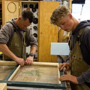 Twee jonge vakmensen werken geconcentreerd aan het plaatsen van glas in een houten kozijn in een werkplaats.