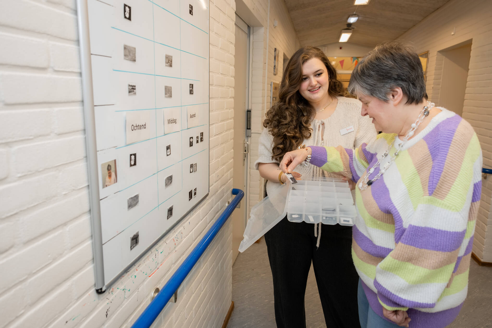Twee begeleiders bespreken rooster op een prikbord in een schoolgang.