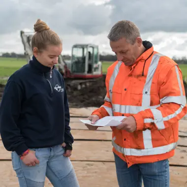 Leerling en instructeur bespreken werkplannen bij grondverzet op een infra-locatie