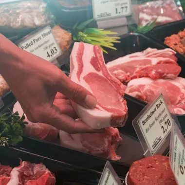 Persoon pakt een stuk vlees bij een toonbank met diverse vleessoorten.