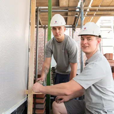 Twee bouwstudenten meten een wand op een bouwplaats met steigers en bakstenen.