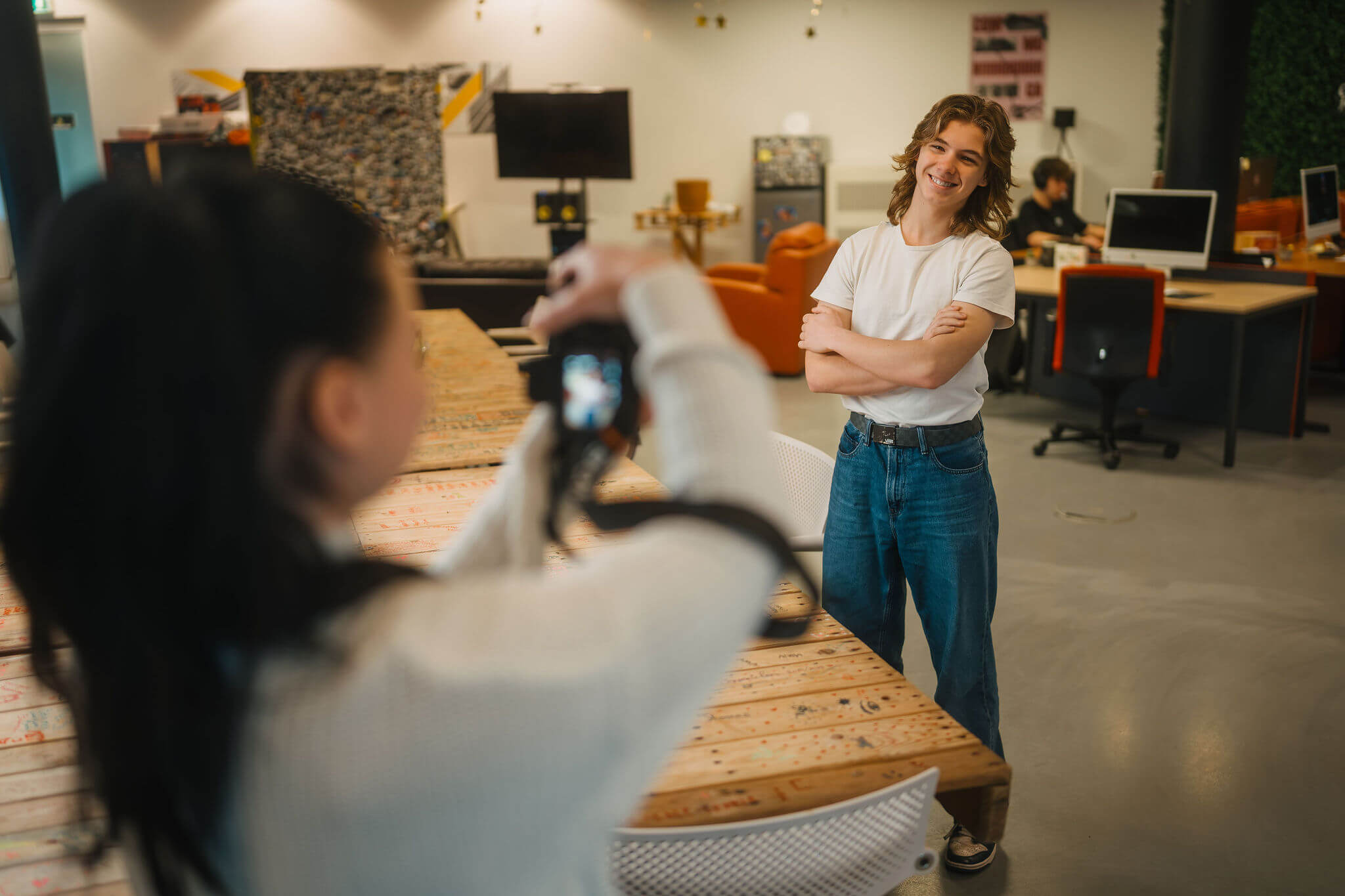 Student poseert bij tafel in creatieve studieomgeving terwijl medestudent foto's maakt met camera