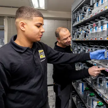 Technische vakdocent begeleidt leerling bij het selecteren van autolak in een werkplaats voor carrosserieherstel