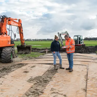 Twee vakmensen bespreken plannen op een bouwterrein met grondverzetmachines en metalen rijplaten.
