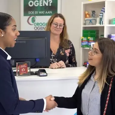 Twee personen schudden handen in een zorg- of medisch-technische omgeving met balie en apparatuur op de achtergrond.