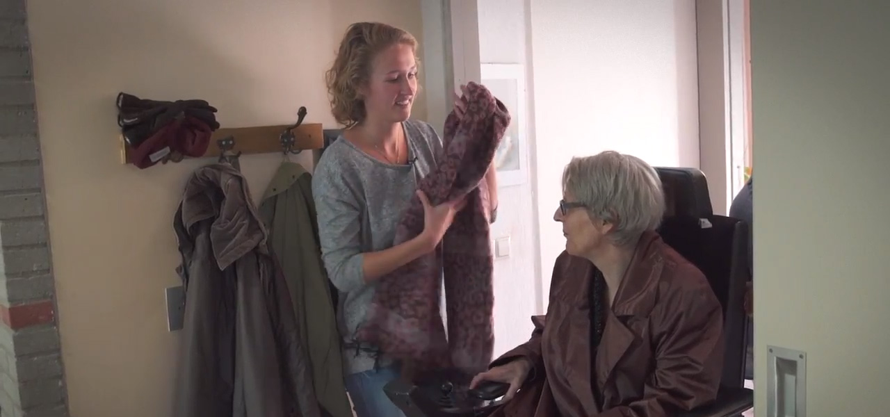 Verzorgende reikt kleding aan zorgvrager in rolstoel tijdens zorgmoment in huiselijke omgeving