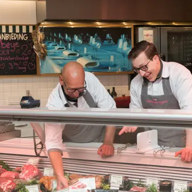 Twee medewerkers helpen klanten bij een vlees- en traiteursbalie met ambachtelijke producten