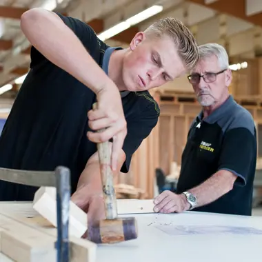 Student oefent timmerwerkzaamheden onder begeleiding in werkplaats met hout en tekeningen