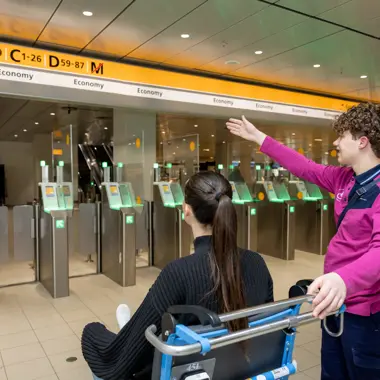 Medewerker begeleidt reiziger in rolstoel bij toegangspoortjes in een openbare vervoersomgeving