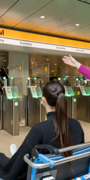 Medewerker begeleidt reiziger in rolstoel bij toegangspoortjes van luchthaven, toont route naar incheckbalie