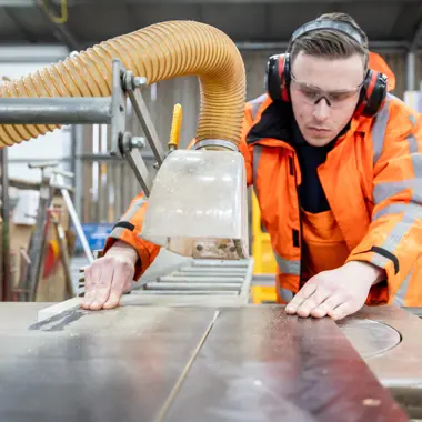 Vakman in oranje veiligheidskleding werkt geconcentreerd met een zaagmachine in een werkplaats voor infra.