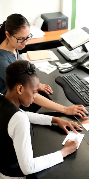 Leerling ontvangt begeleiding achter de balie in een horecagelegenheid, werkend met computer en papieren.