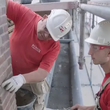 Twee bouwvakkers plaatsen bakstenen aan een gevel op een steiger tijdens de bouw van een woning
