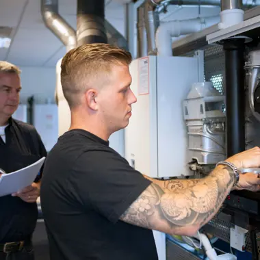 Monteur werktuigkundige installaties voert onderhoud uit aan een cv-ketel onder begeleiding van een collega.