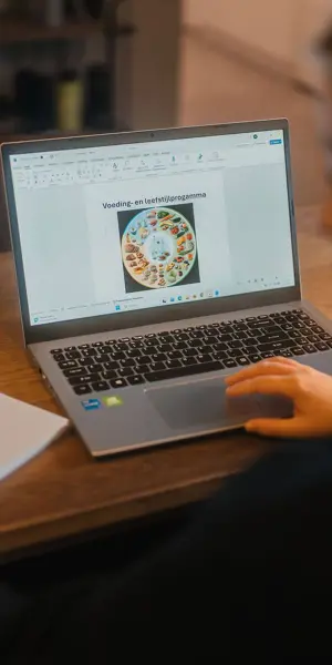 Student werkt aan opdracht over voeding en leefstijlleer op laptop met notitieboek op tafel.
