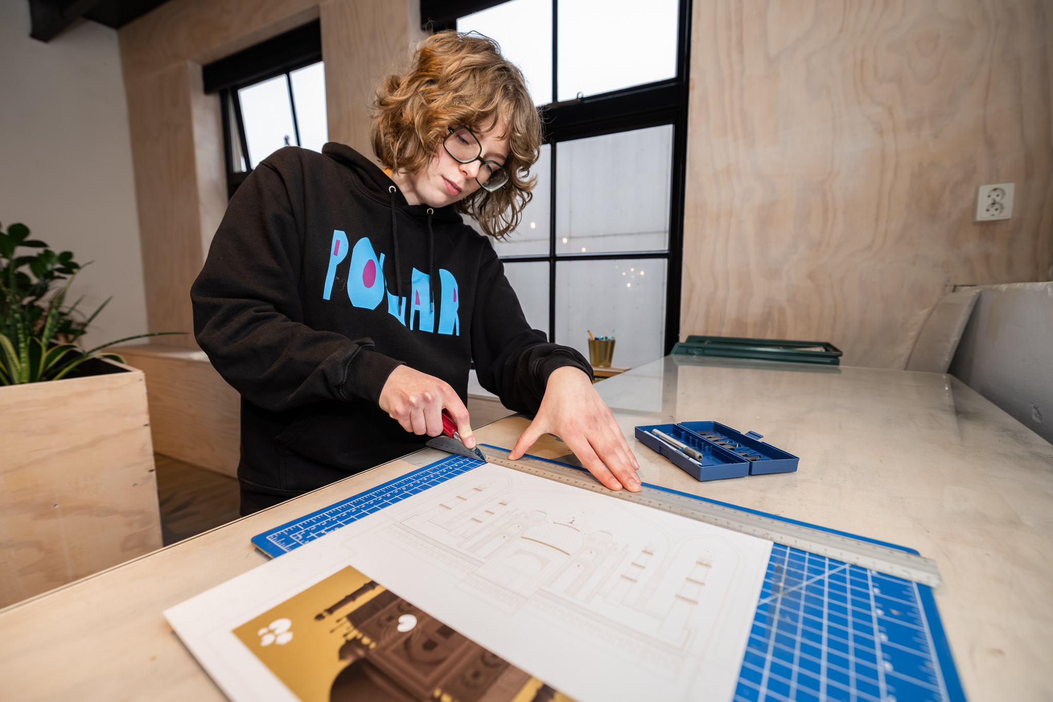 Student gebruikt snijmat en liniaal om nauwkeurig te werken aan ontwerpplaat met gereedschap op tafel