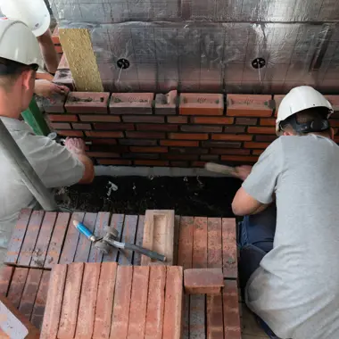 Twee bouwvakkers met helmen metselen een bakstenen muur, bezig met het vormen van een gevel.