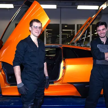 Twee technici in werkkleding werken aan een oranje sportauto met open deuren in een garageomgeving