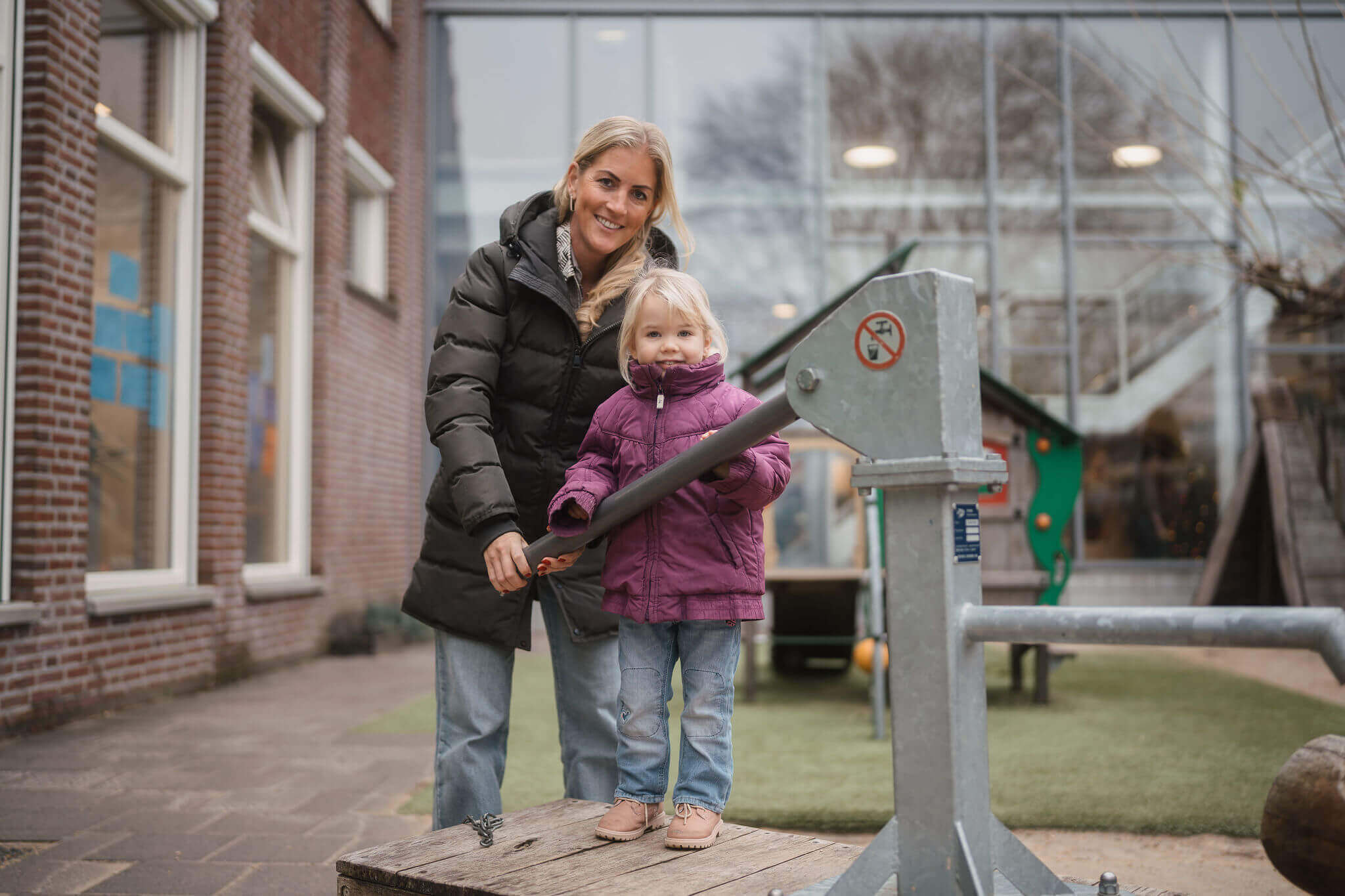 Begeleider toont jong kind hoe waterpomp op schoolplein te gebruiken