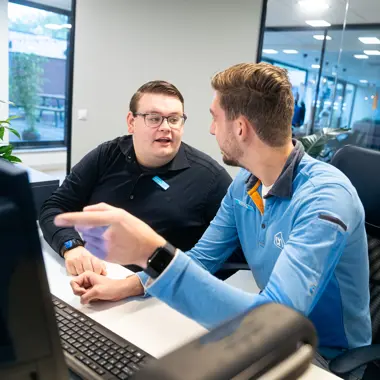 Twee medewerkers bespreken werkprocessen achter een computersysteem in een zakelijke omgeving.