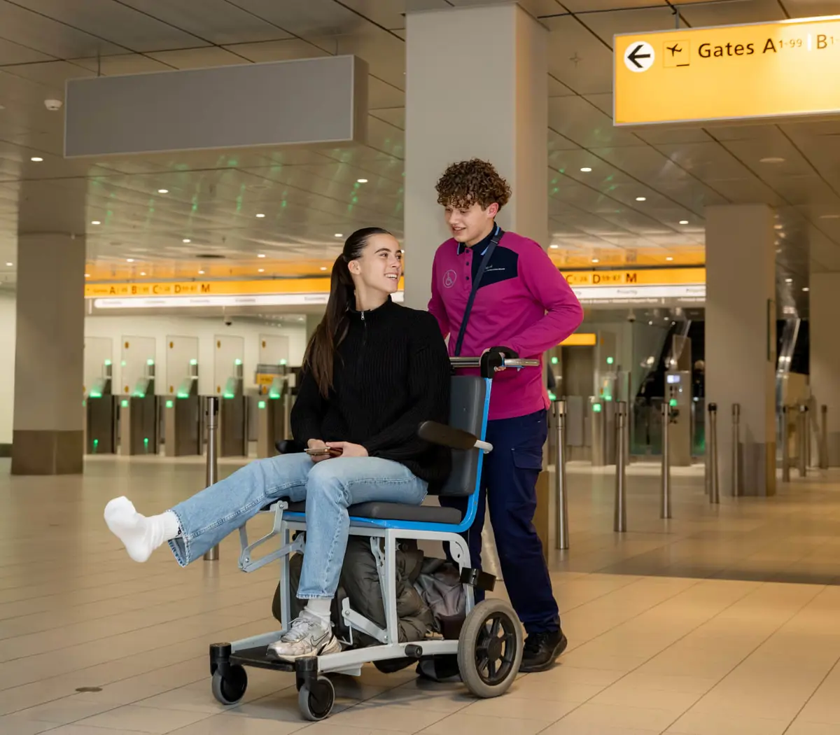 Een luchtvaartdienstverlener begeleidt een reiziger in rolstoel in een luchthavenhal richting gate A-B-C-D.