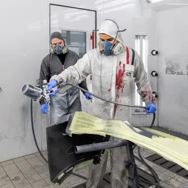 Technici voeren spuitwerk uit op een auto-onderdeel in een professionele werkplaats.