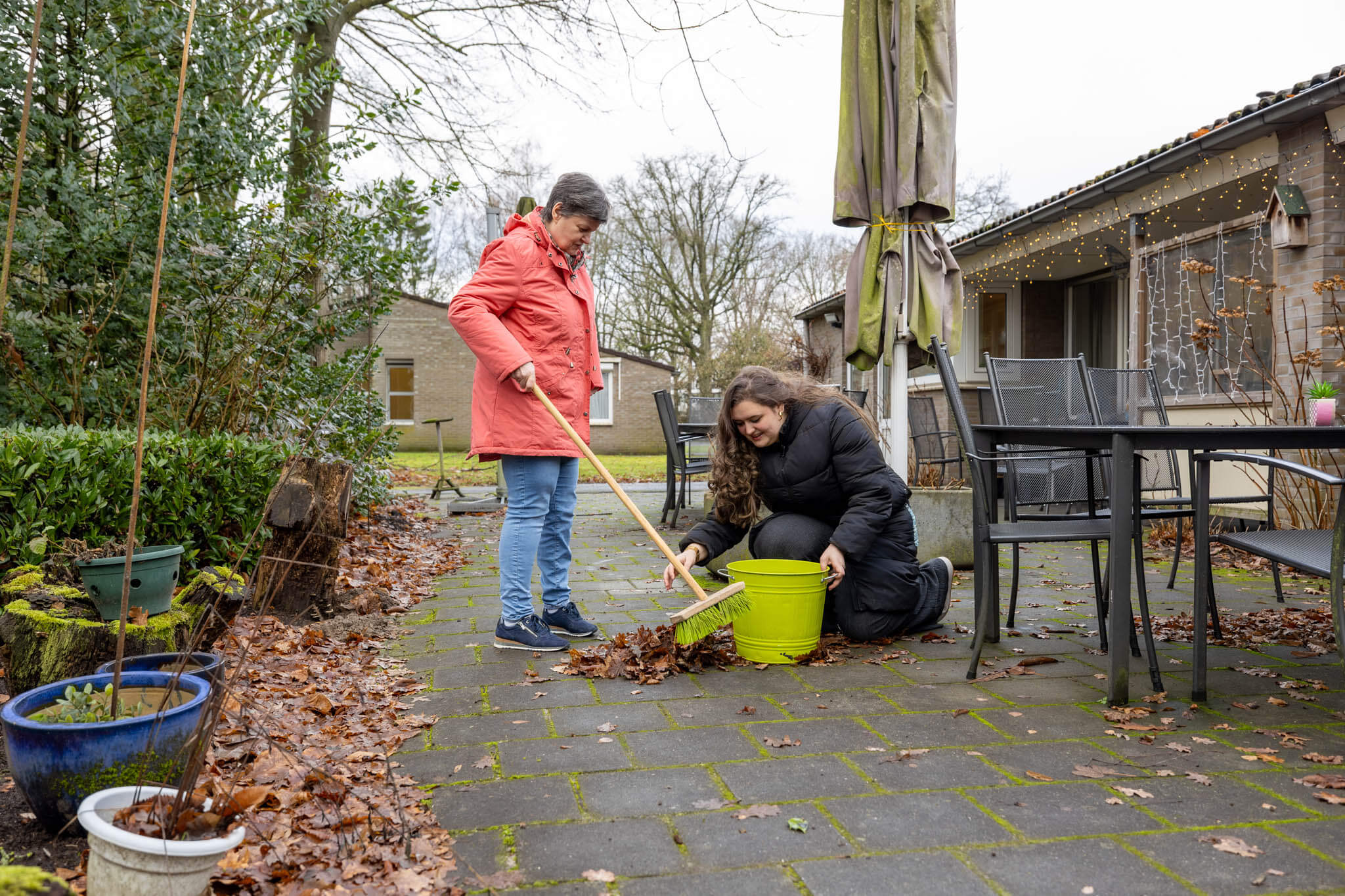 Twee personen ruimen bladeren op in een tuin met gereedschap, gericht op samenwerken en buitenwerk