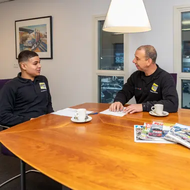 Twee medewerkers bespreken autocarrosserie en onderhoud aan een tafel met brochures en koffie.
