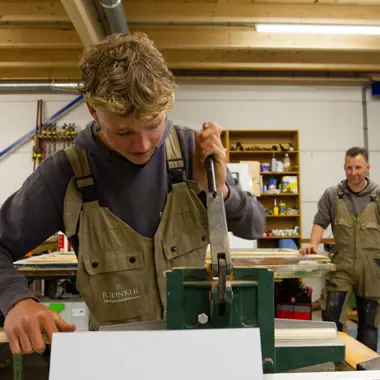 Leerling werkt geconcentreerd in een werkplaats aan houtbewerking, begeleider kijkt toe vanuit de achtergrond