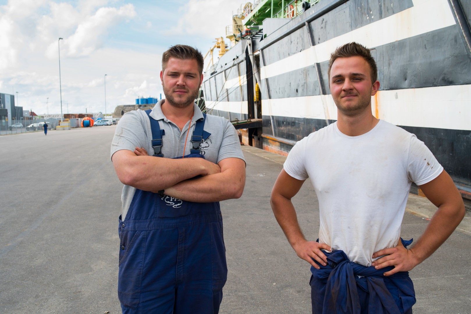 Twee jongeren in werkkleding op een kade naast een maritiem schip, klaar voor werk aan boord.