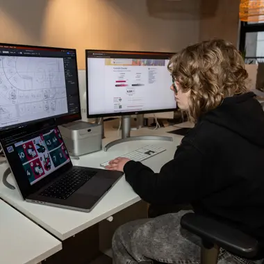 Student werkt geconcentreerd aan ontwerp- en communicatieopdrachten op meerdere schermen binnen creatieve mediaomgeving