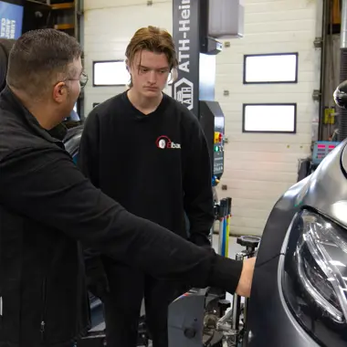 Docent legt aan leerling uit hoe onderdelen van een auto gecontroleerd en gerepareerd worden in een werkplaats